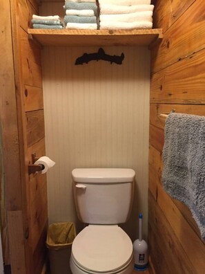 Ein kleines Badezimmer mit Holzverkleidung, ausgestattet mit Toilette, Ablagefläche über der Toilette mit gefalteten Handtüchern und einem Handtuch an der Wand.