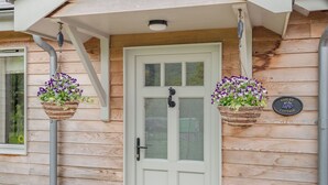Ein hölzernes Häuschen mit weißer Tür, zwei hängende Blumenkörbe und einem Schild mit der Aufschrift 'VIOLET COTTAGE'.