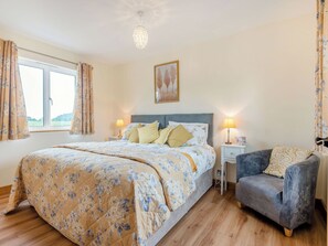 Ein Schlafzimmer mit Bett, Nachttischlampen, einem Sessel, einem Fenster mit Vorhängen und einem Bild an der Wand.