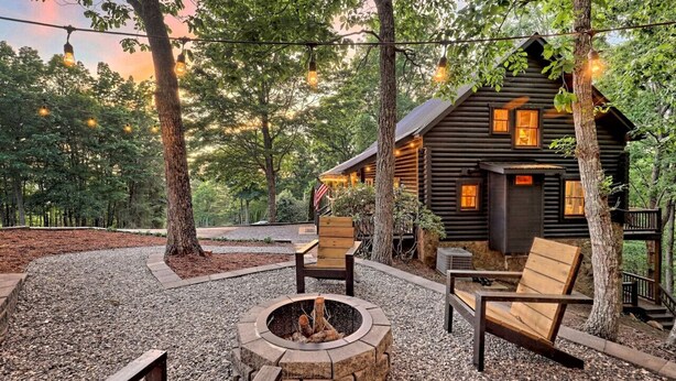 Una baita rustica con un braciere e posti a sedere all'aperto, immersa tra gli alberi e illuminata da luci decorative.