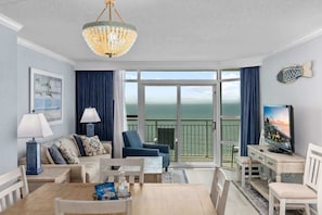 Ein Zimmer mit einer Couch, Sesseln, einem Esstisch, einem Fernseher und einem Balkon mit Blick.