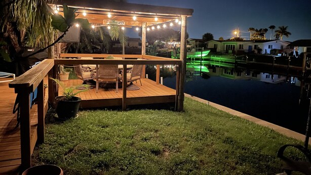 A wooden deck with a pergola and string lights, overlooking a calm water body with boats, surrounded by greenery. - Amazing waterfront home on Palma Sola Bay, a bicycle ride to  Anna Maria Island. (Bradenton)