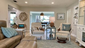 A spacious living room with a beige leather sofa, a matching armchair, and a small round table with a lamp. A large window provides natural light, and the room features a decorative wall piece and a framed picture. - Amazing waterfront home on Palma Sola Bay, a bicycle ride to  Anna Maria Island. (Bradenton)