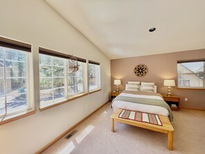Une chambre à coucher avec un grand lit, deux tables de chevet avec des lampes, une horloge murale et une fenêtre donnant sur des arbres.