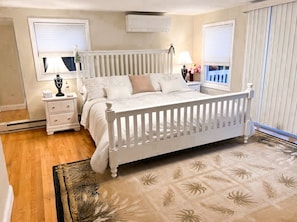 Une chambre à coucher avec un lit blanc, une table de chevet, une lampe et une fenêtre avec des stores.