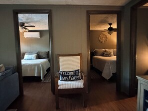 Habitación con dos camas, una silla y un ventilador de techo. Se ve un letrero que dice 'Welcome to the Lake House'.