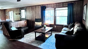A living room with a wooden floor, a sofa, a chair, a coffee table, and a window with curtains. - Cozy Cabin with Screened Porch and Fishing Dock on Lake D'Arbonne (Downsville)