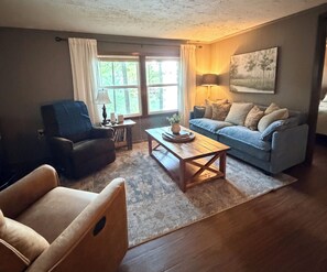 Una sala de estar con sofá, sillón, mesa de centro y un cuadro en la pared.