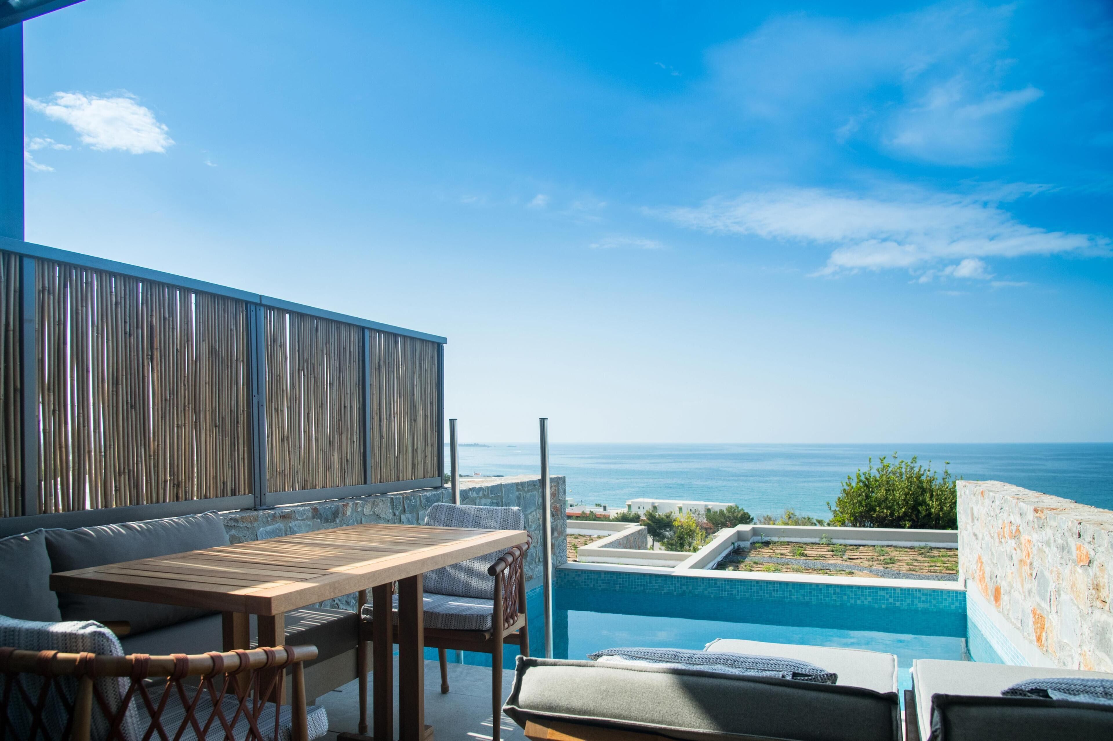 A terrace with a wooden table and chairs, a pool, and a view of the sea.
