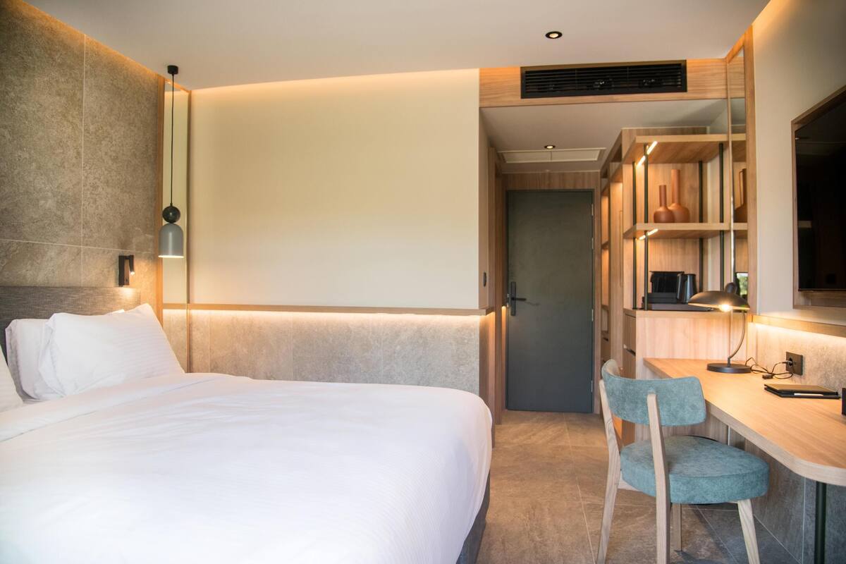 A modern hotel room with a large bed, a desk with a chair, and a built-in wardrobe.