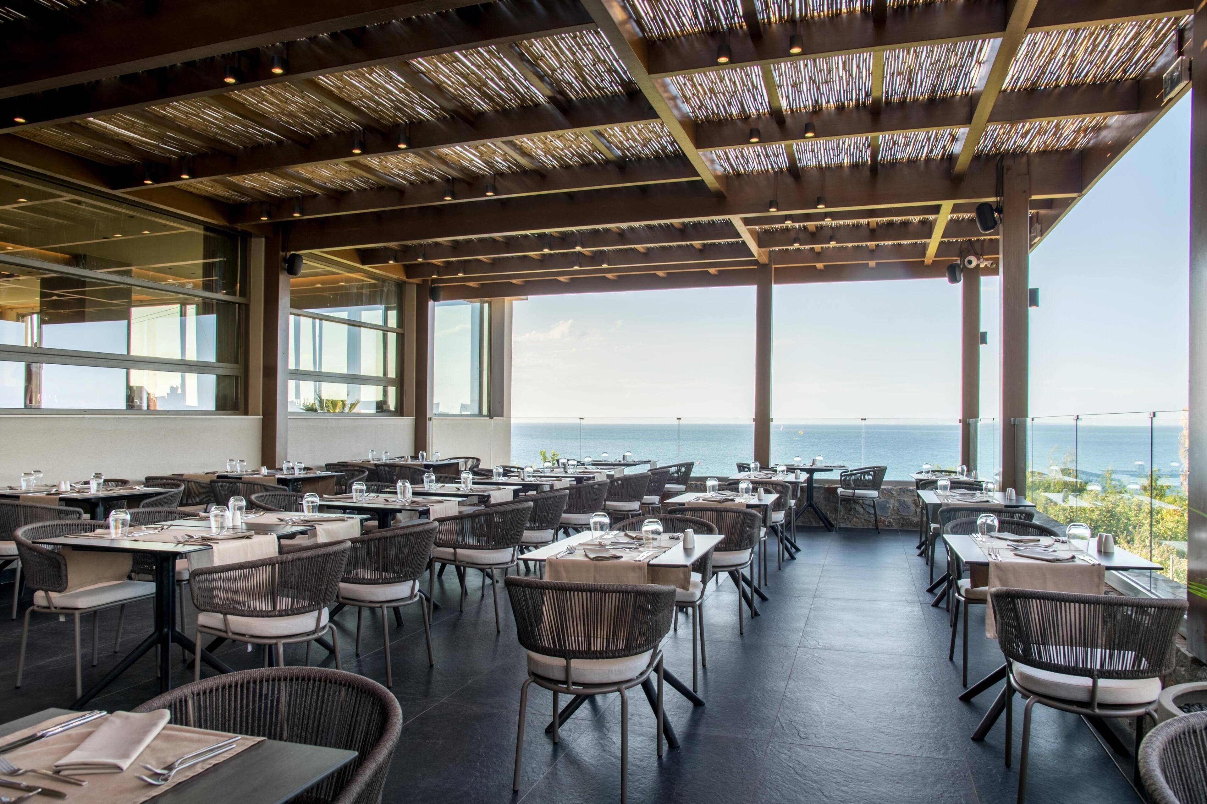 A spacious outdoor dining area with tables and chairs, large windows, and a view of the sea.