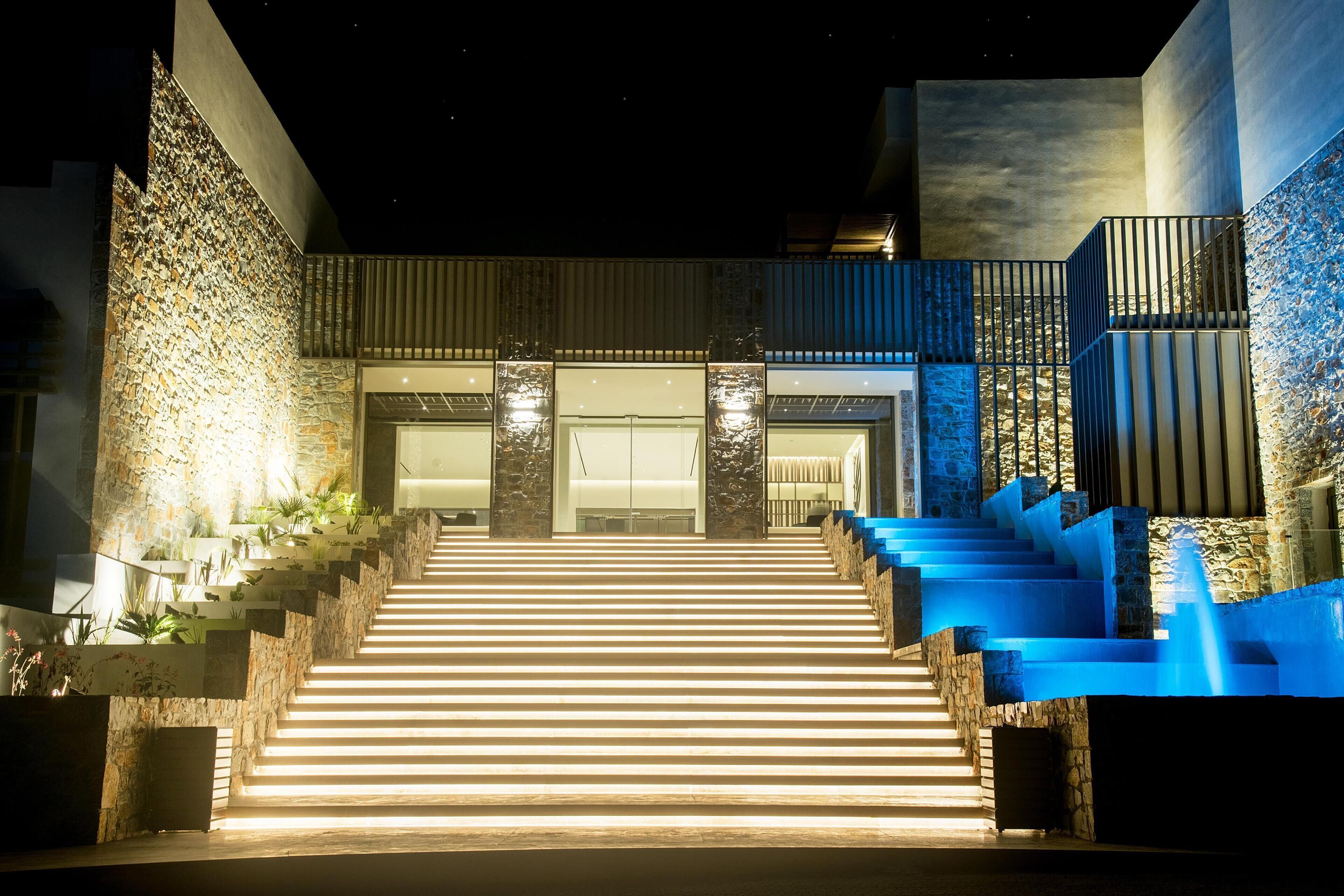 A nighttime view of a hotel entrance with a staircase leading up to a gated door, surrounded by stone walls and blue lighting.