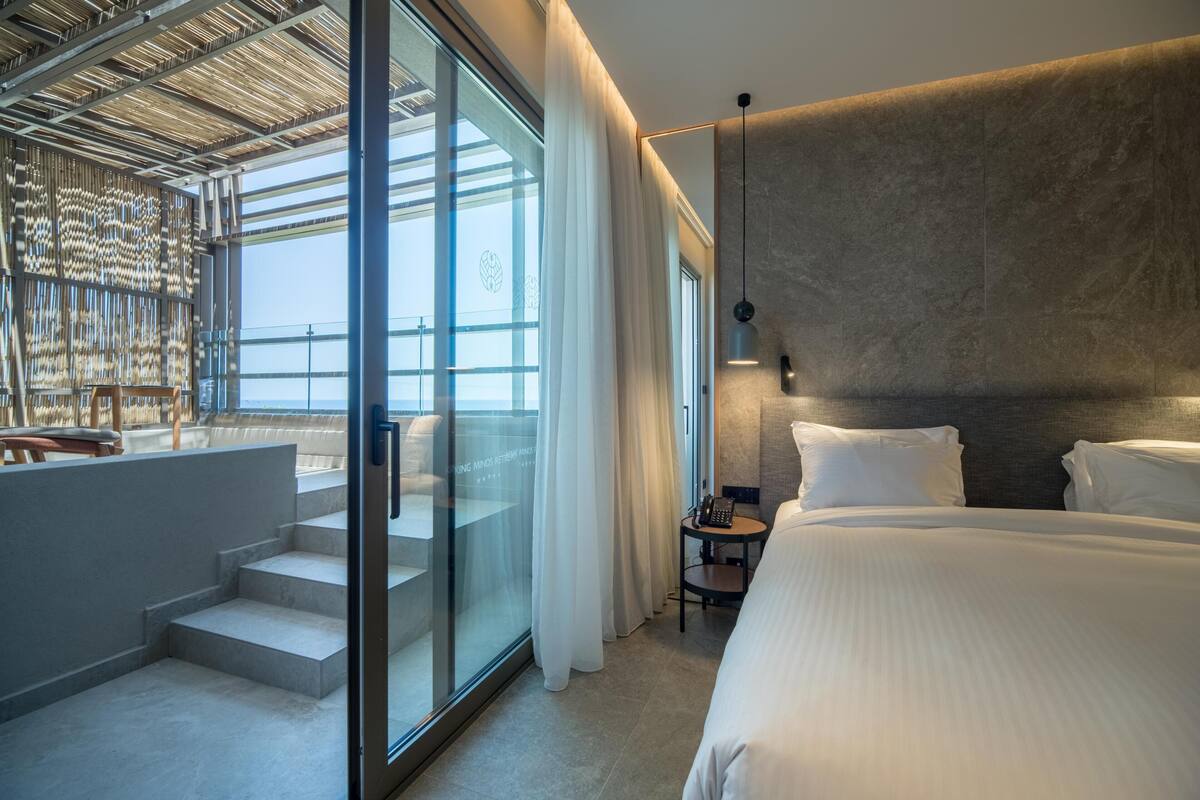 A modern hotel room with a large bed, a glass wall with a view, and a staircase leading to a balcony.