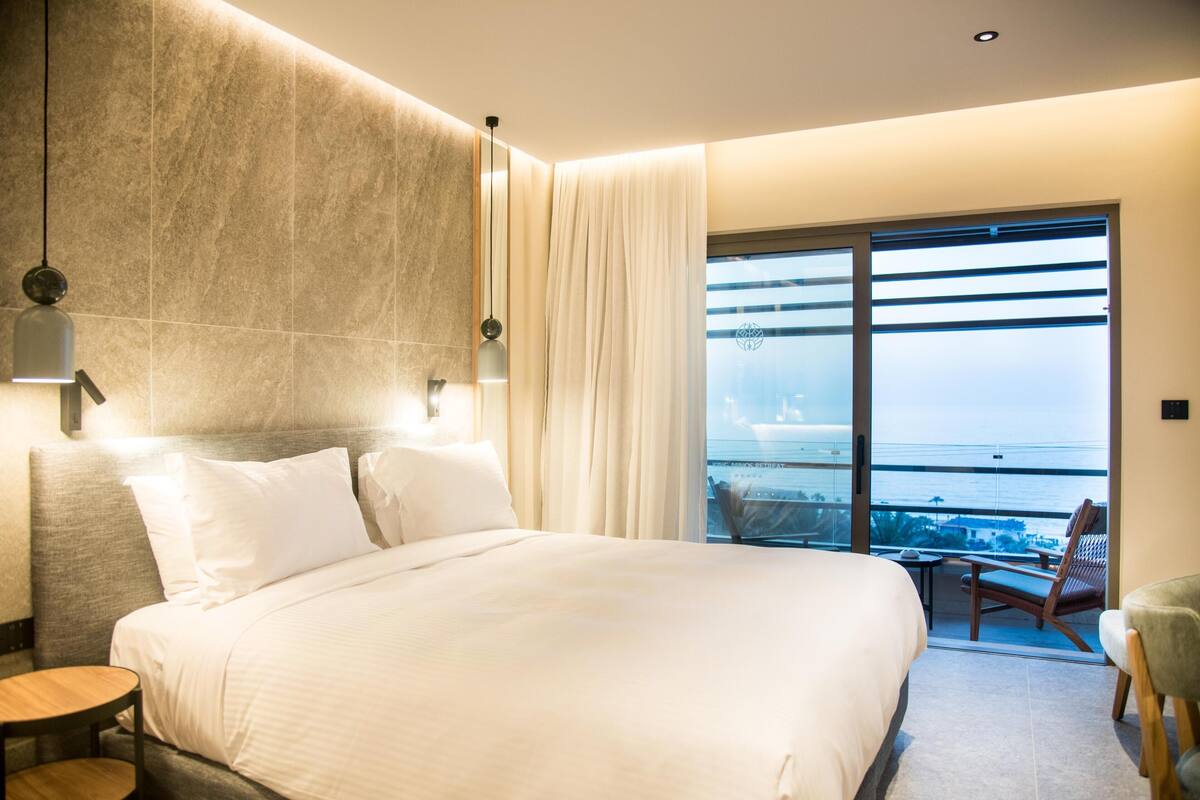 A modern hotel room with a large bed, a view of the sea through floor-to-ceiling windows, and a balcony with a chair.