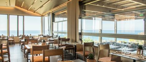 A restaurant with tables set for dining, large windows offering a view of the sea, and a modern interior design.