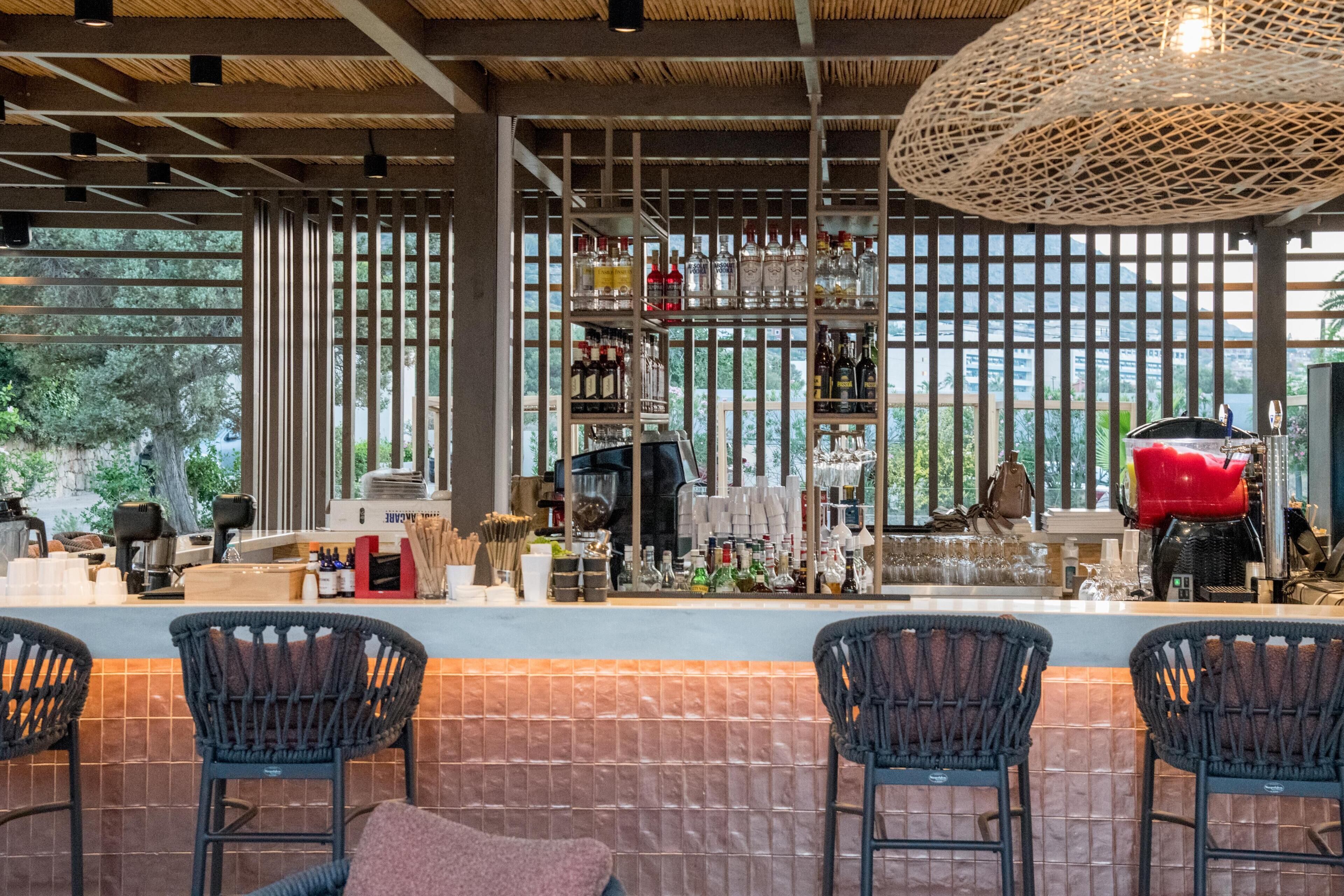 A bar area with wicker chairs, a counter with various bottles, and a view of trees through large windows.