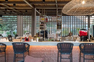 Un espace bar avec des fauteuils en osier, un comptoir où sont exposées différentes bouteilles, et une vue sur les arbres à travers de grandes fenêtres.