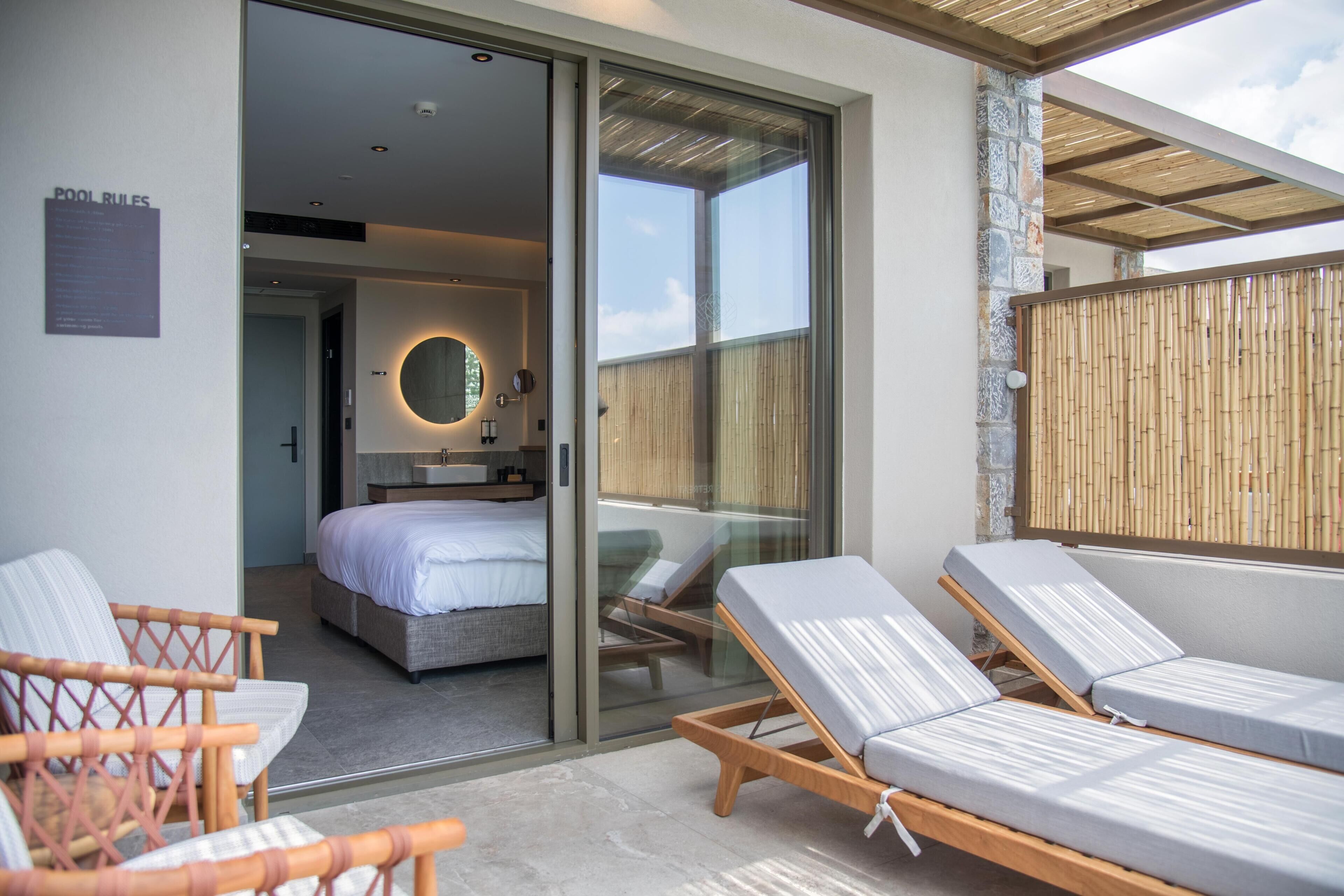 Una habitación de hotel moderna con puerta de vidrio que da a un balcón, una cama, un baño y dos sillones.