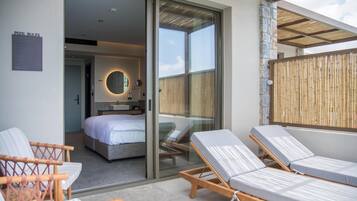 Una habitación de hotel moderna con puerta de vidrio que da a un balcón, una cama, un baño y dos sillones.