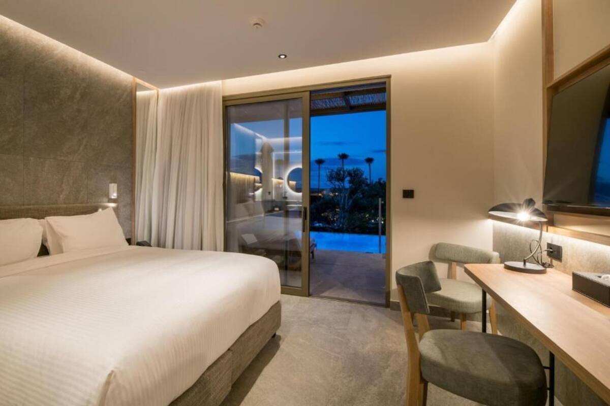 A modern hotel room with a large bed, a desk, and a glass door leading to a balcony.