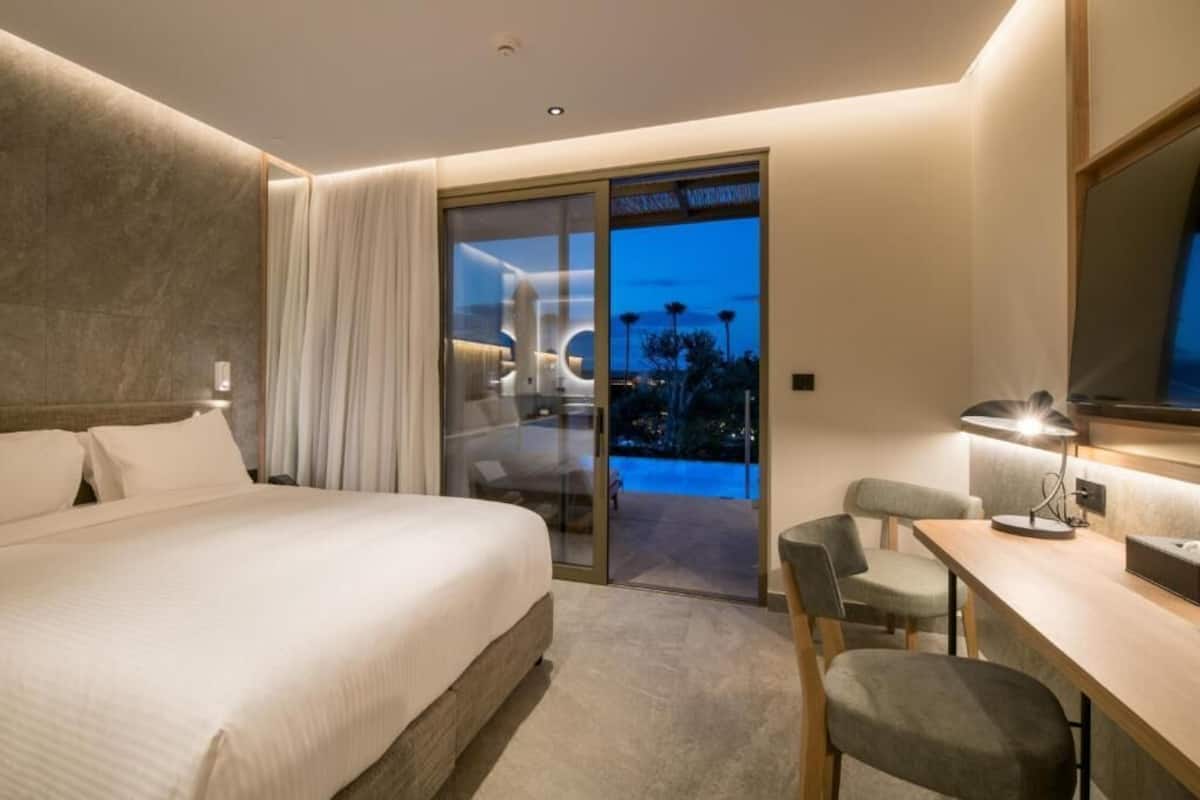 A modern hotel room with a large bed, a desk, and a glass door leading to a balcony.