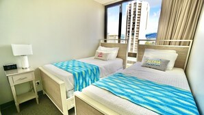 A hotel room with two beds, a nightstand with a lamp, and a view of a cityscape through large windows. - Modern 2BR w/Diamond Head view, beach, FREE parking, king bed, pool, gym,& more! (Honolulu)