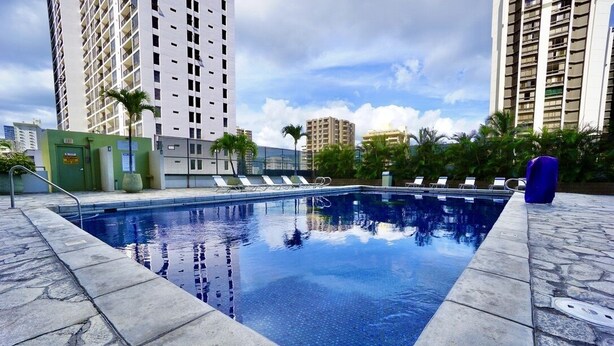 A rectangular swimming pool with a stone border, surrounded by lounge chairs and palm trees, with tall buildings in the background. - Modern 2BR w/Diamond Head view, beach, FREE parking, king bed, pool, gym,& more! (Honolulu)