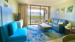 A modern living room with a blue sofa, a grey sofa with yellow cushions, a coffee table, and a TV on a stand. There is a large window with a view of a mountain and a balcony with a table and chairs. - Modern 2BR w/Diamond Head view, beach, FREE parking, king bed, pool, gym,& more! (Honolulu)