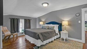 Une chambre à coucher avec un lit, deux lampes de chevet, une chaise en bois, une fenêtre avec des rideaux et un tapis à motifs.