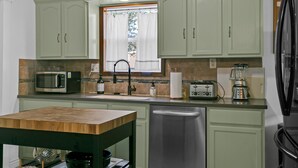 Une cuisine avec des meubles de couleur vert clair, un plan de travail en bois, un lave-vaisselle en acier inoxydable, un four à micro-ondes, un grille-pain et une fenêtre avec des rideaux.