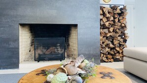Een woonkamer met een open haard, een ronde houten salontafel met decoratieve objecten, een beige bank en een stap hout tegen de muur.