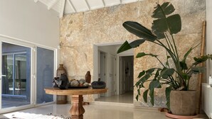 Een ruime kamer met een marmeren wand, een ronde houten tafel met decoratieve objecten, en een grote potplant.