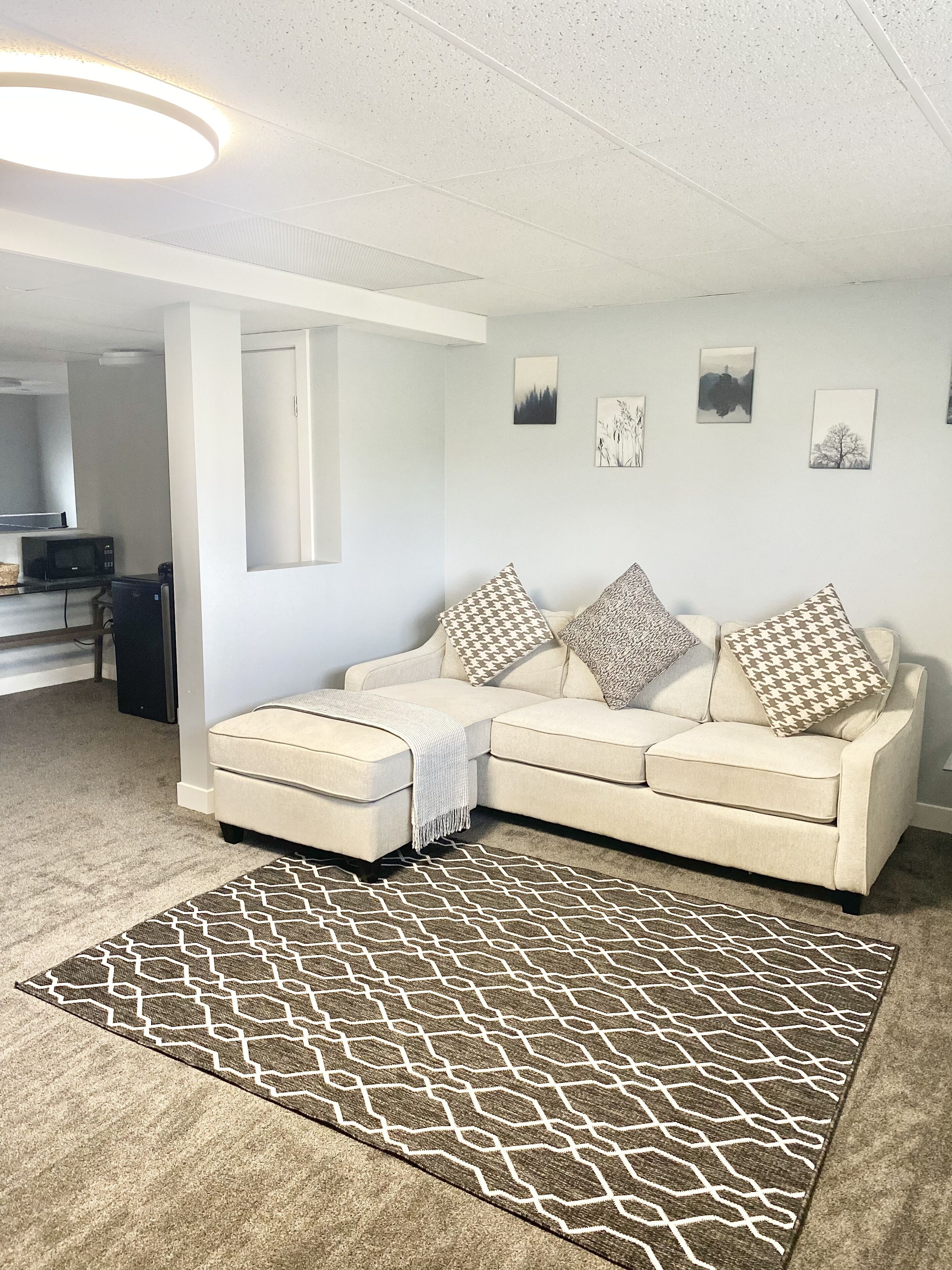 Una sala de estar con un sofá seccional color beige, almohadones estampados, un tapete geométrico y una zona de cocina al fondo.