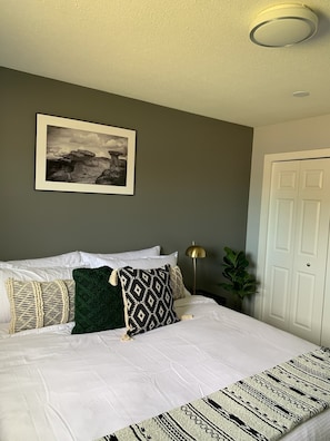 Un letto rifatto con lenzuola bianche e cuscini decorati, un quadro appeso al muro e una porta sulla destra.