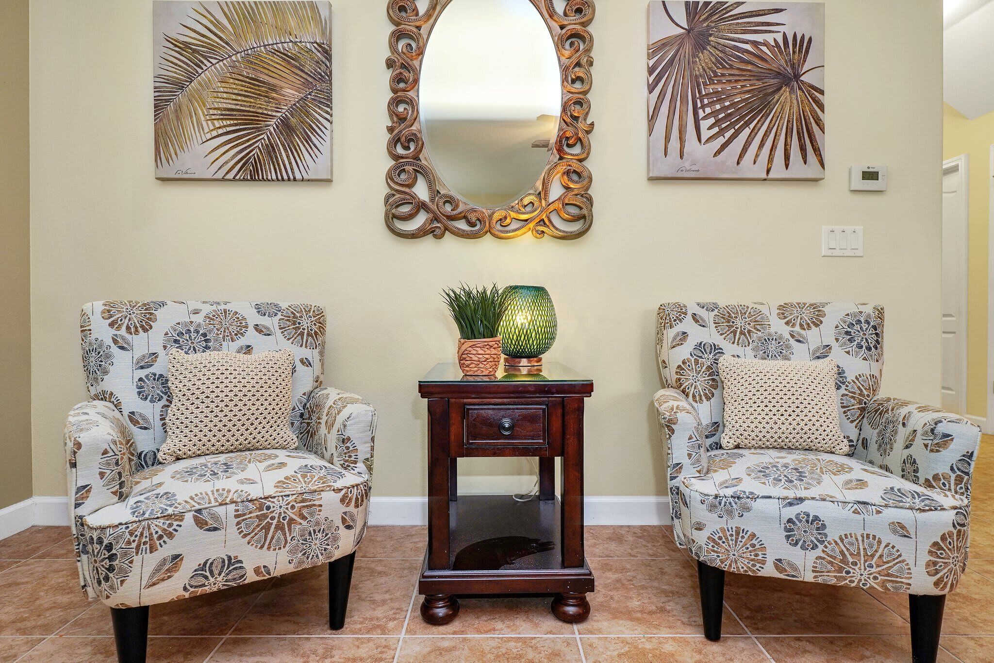 Deux fauteuils à motifs, séparés par une petite table d’appoint en bois, un miroir décoratif au-dessus, et deux œuvres d’art accrochées au mur.