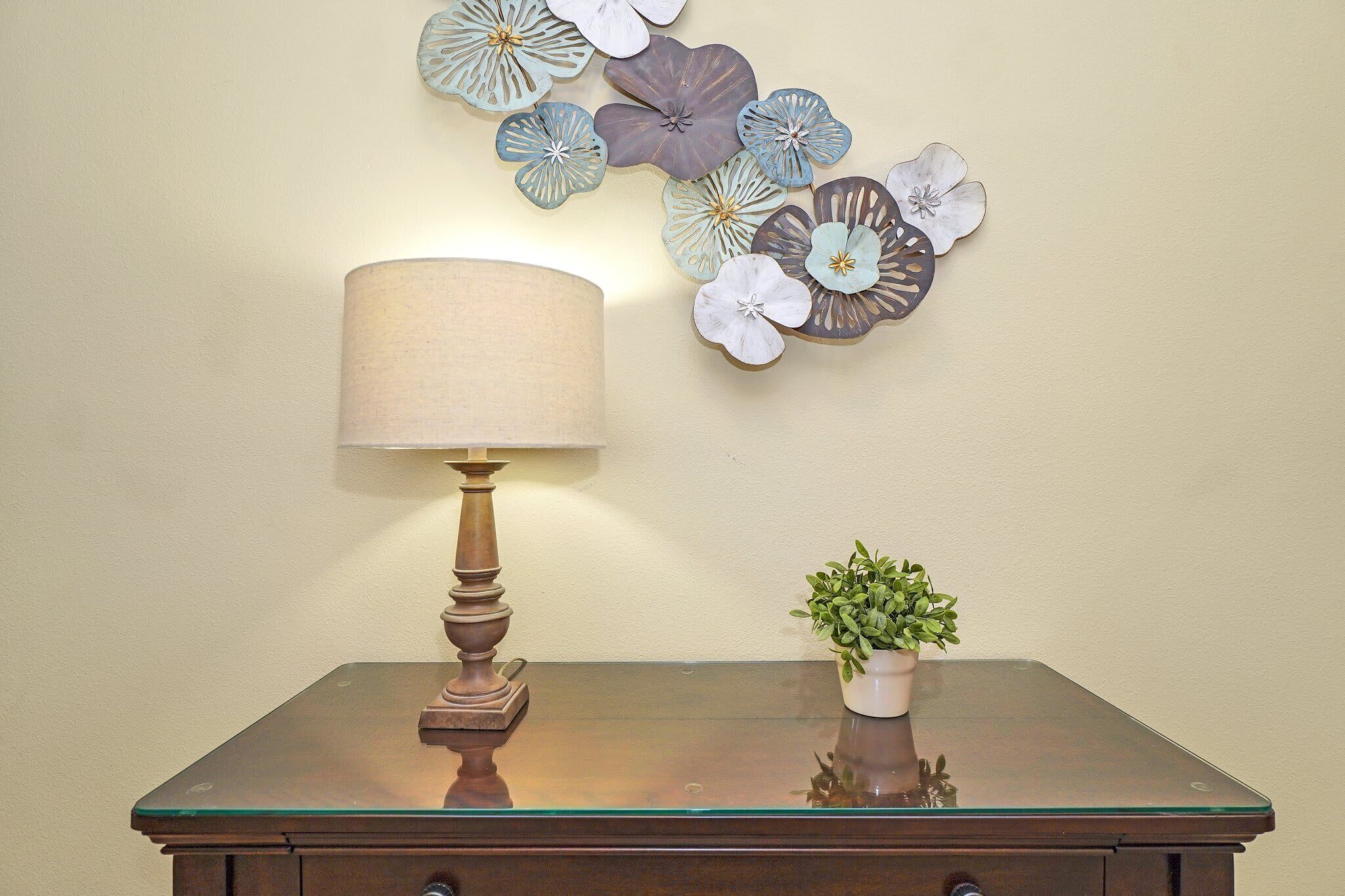 Une table avec une lampe et une plante en pot, placée devant un mur orné d’un décor floral.