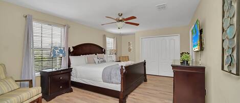 Une chambre à coucher comprenant un lit, une table de chevet, une commode, une chaise, un ventilateur de plafond et une porte.