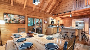Interior de una cabaña de madera con una mesa de comedor preparada para seis personas, zona de cocina con refrigerador y un ventanal con vistas al bosque.