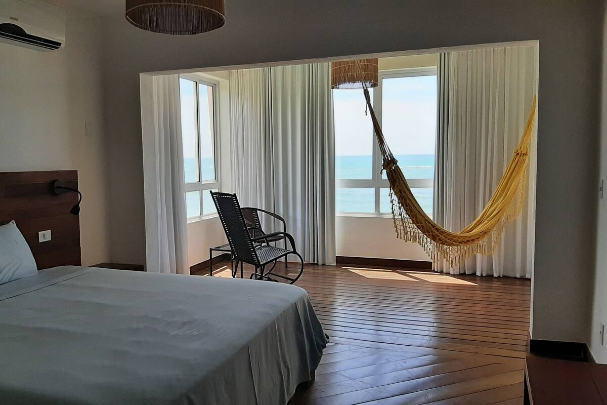 A hotel room with a bed, a chair, a hammock, and a view of the sea.