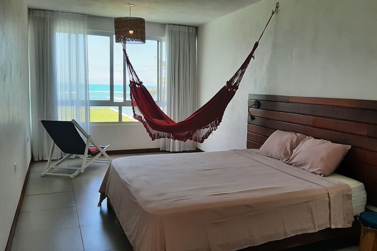 A bedroom with a bed, a hammock, a chair, and a view of the sea.
