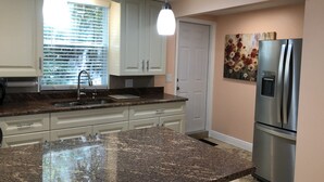 A kitchen with granite countertops, white cabinets, a stainless steel refrigerator, and pendant lighting.