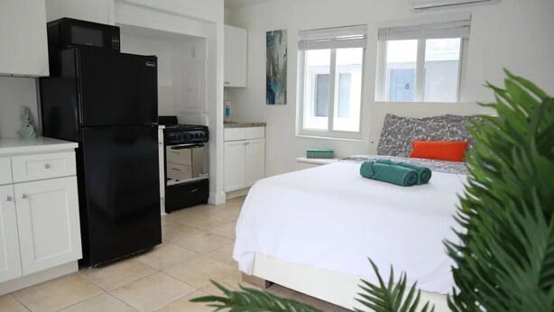 A bedroom with a bed, a window, a kitchen area with a refrigerator, and a small table.