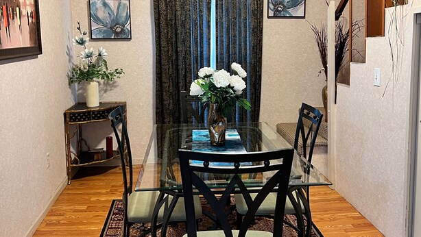 A dining area with a glass table, four chairs, a vase with flowers, a patterned rug, and two framed pictures on the walls. - Comfy Clean & Cute (Marion)