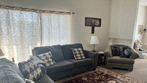 A living room with a grey sofa, a patterned armchair, a red and beige rug, and a wall-mounted lamp. - Comfy Clean & Cute (Marion)