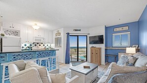 A living room with a sofa, coffee table, and a kitchen area with white cabinets and a blue backsplash. - Gulf Shores Plantation-Dunes 5208 (Gulf Shores)