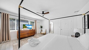 A hotel room with a large bed, a TV, a ceiling fan, a wooden dresser, a chair, and a window with curtains. - Newly Remodeled Home with Spacious Backyard! (Fort Lauderdale)