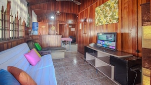 Una habitación acogedora con detalles de madera, un sofá con cojines coloridos, un televisor sobre un mueble bajo y una zona de comedor al fondo.