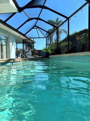 A swimming pool with a glass ceiling structure, clear blue water, and a view of palm trees and a house in the background.