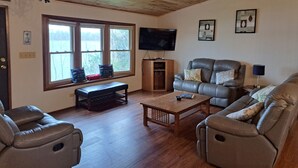 Ein Wohnzimmer mit Holzbalkendecke, Ledersofas, einem Couchtisch aus Holz, einer Bank und einem Fernseher auf einem Holzständer.
