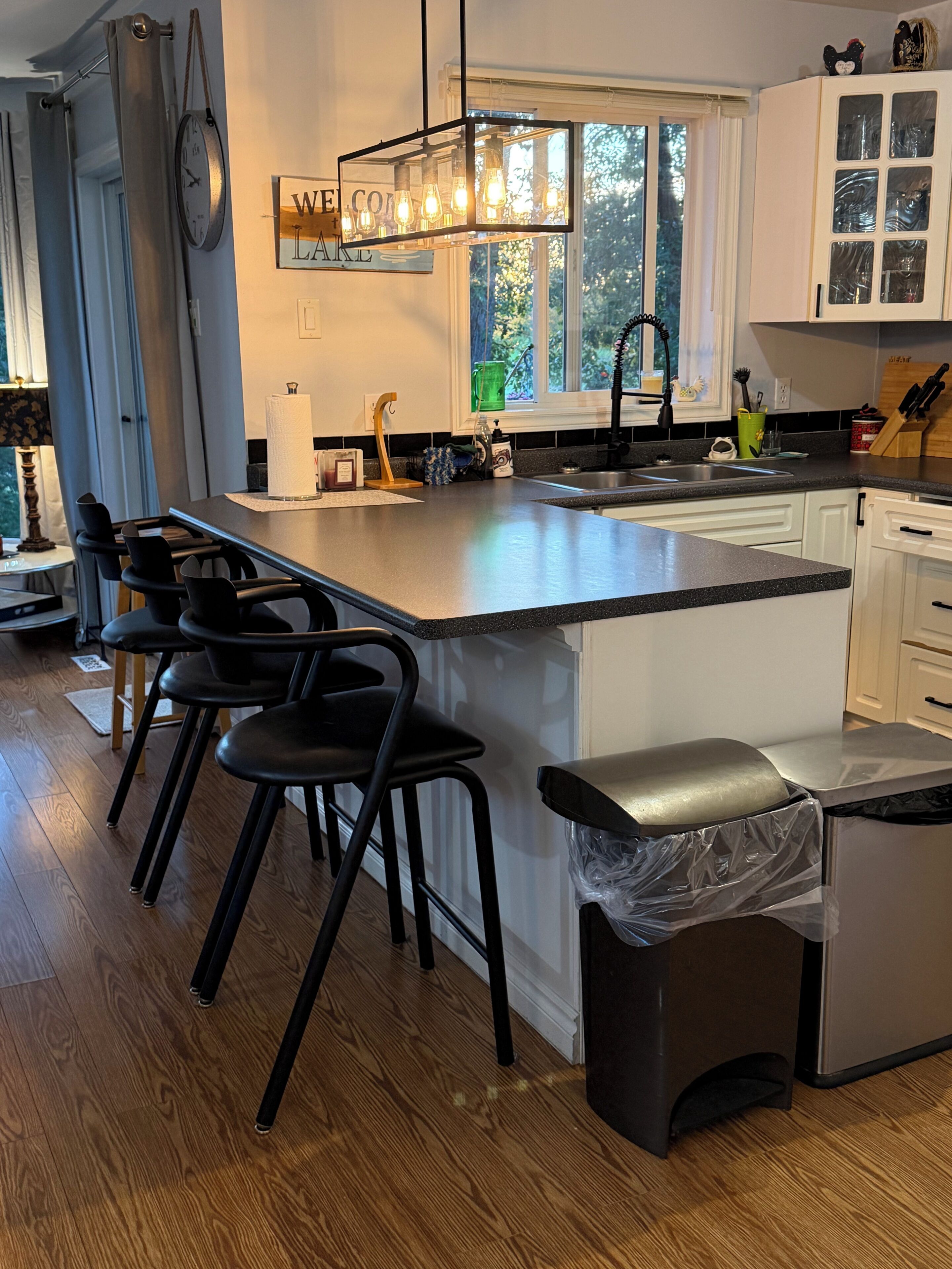 A modern kitchen with a central island, bar stools, a clock, and a window with a view of trees.